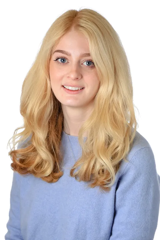 A smiling young woman with long blonde and light brown wavy hair wears a light blue sweater.