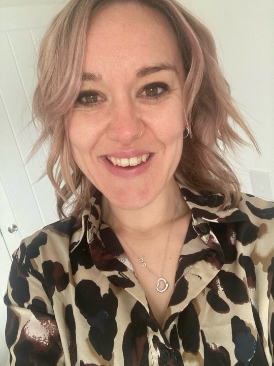 A smiling woman with light brown hair and pink highlights wears a leopard print blouse and a silver heart necklace.