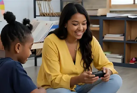 A mother and a child sitting on the floor, smiling and looking at a video game controller. The mother is holding the controller while the child watches with a happy expression.